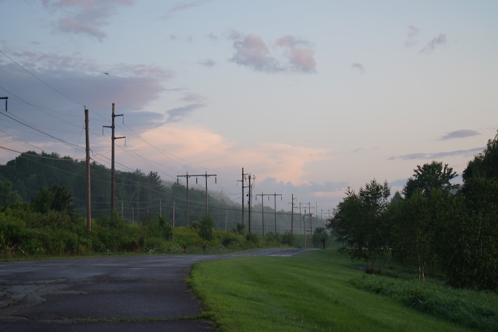 Photo depicting a blue sky with some purple and pink clouds mainly focused on the left side of the photo, there is a road and next to it some grass and shrubs. there are power lines following the road.