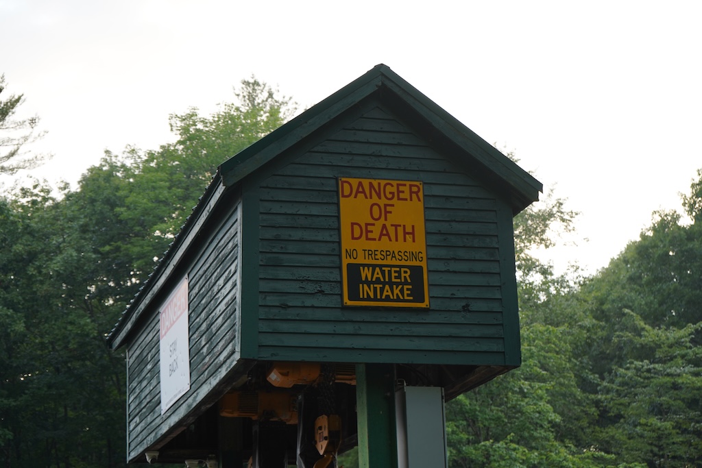 Photo of some sort of wooden structure painted a forest green, in the middle of the structure there is a sign saying "DANGER OF DEATH NO TRESPASSING WATER INTAKE", in the background there are some trees.
