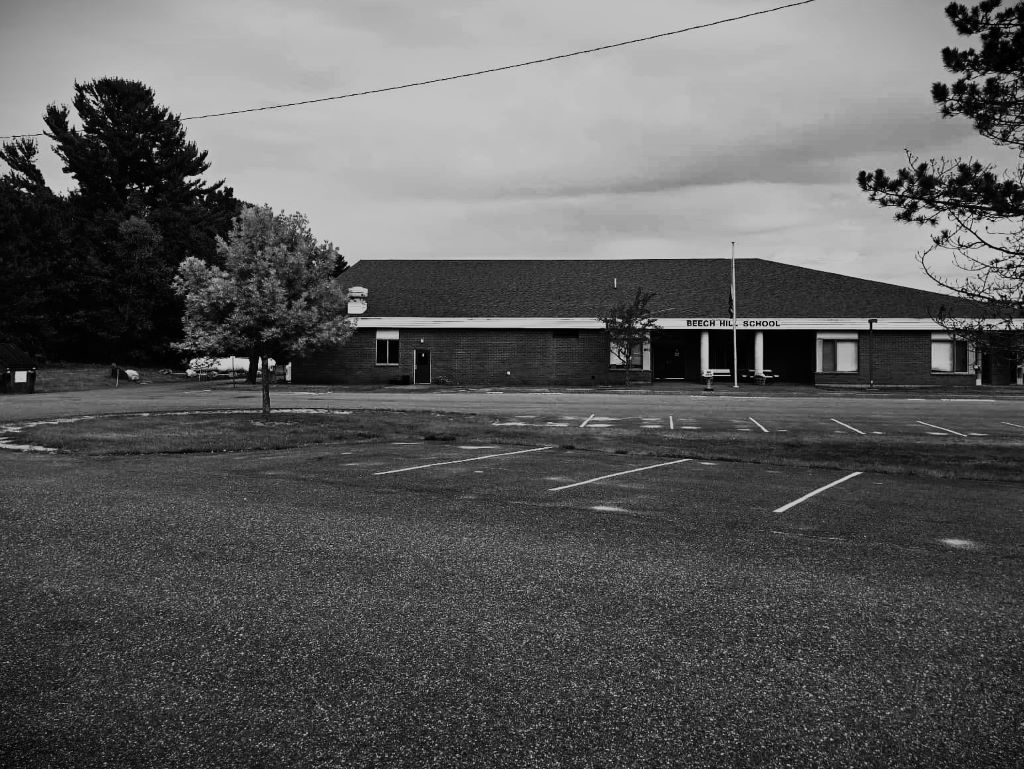 A black and white deserted elementary-middle school parking lot. Gloomy clouds roll overhead the empty front steps of the school.