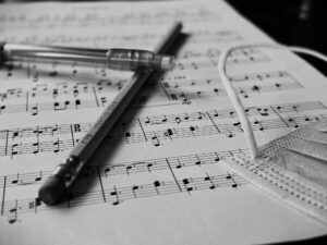 A black and white sheet of piano music. An untouched and unsharpened pencil rests alongside a COVID mask and mechanical pencil, casting shadows of work incomplete.