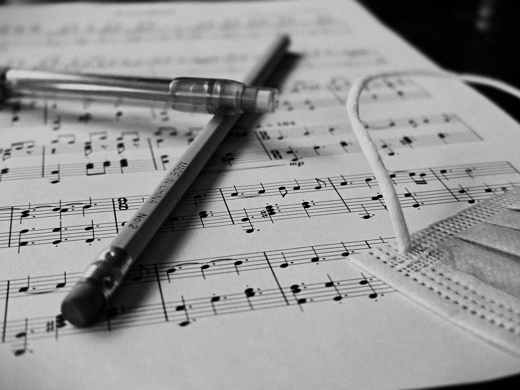 A black and white sheet of piano music. An untouched and unsharpened pencil rests alongside a COVID mask and mechanical pencil, casting shadows of work incomplete.