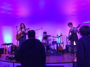 The singer/acoustic guitarist, electric guitarist, and drummer of Milk St performing on a small stage with purple backlighting. There's a few people standing near the front of the stage.