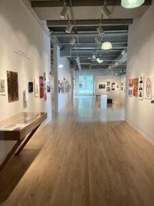 A head-on photo of the Lord Hall gallery's main isle, walls lined with all kinds of paintings and woodblock prints, and a couple of small sculptures on stands in the distance. The place is lit up with studio lights and sunlight from a window in the back.