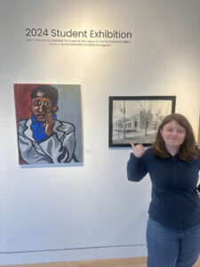 A picture of me, a pudgy 5' 3" redheaded woman in a blue sweatshirt and jeans, confidently pointing backwards at my painting on a wall in the gallery. The painting is a copy of Alice Neel's portrait of social critic, teacher, and author Harold Cruse, which I painted as part of a class project.
