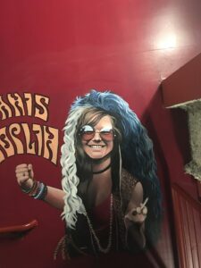 A mural of a smiling Janis Joplin, complete with big 1960s shades and plenty of beaded bracelets, on a red wall by a stairwell.