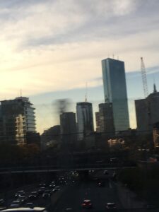 A picture of Boston at sunset, a backdrop of golden-peach clouds contrasting with the severe silhouettes of office high-rises. A couple of streets can be seen below, chaotic as ever, with many cars scrambling to get where they're going in one piece.