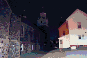 A cobblestone building at night with a large cathedral spire rising up into the darkness. To the right of the building is a bright yellowish-pink residential building. Image has been heavily compressed.