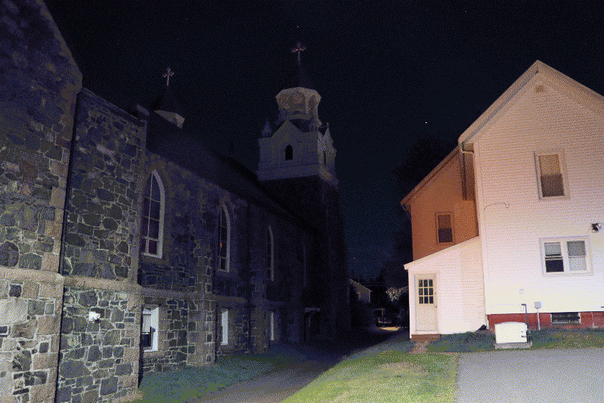 A cobblestone building at night with a large cathedral spire rising up into the darkness. To the right of the building is a bright yellowish-pink residential building. Image has been heavily compressed.
