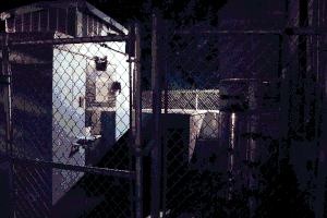 A series of mechanical boxes and pipes behind a building. Objects are viewed from behind a mesh fence. Image has been heavily compressed.
