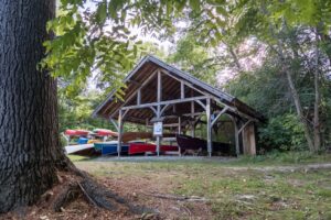 Alongside a trail in a shaded area, a large open-air structure houses a variety of canoes.